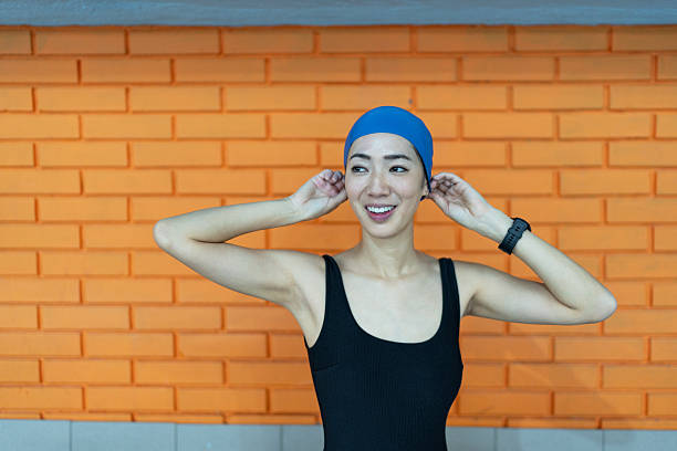 Young adult woman swimmer wearing a blue swimming cap and black swimsuit, smiling while adjusting her cap against an orange brick wall, preparing for a refreshing swim
