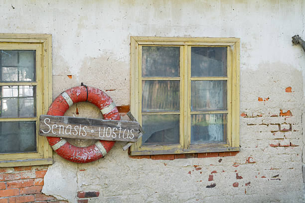 Detailed shot of an aged building's facade, featuring two grimy wooden-framed windows with peeling yellow paint. A heavily weathered red and white lifebuoy hangs between them, adorned with a rustic wooden sign that reads Senasis Uostas (Old Harbor). The wall itself shows a textured mix of crumbling off-white plaster revealing distressed red bricks underneath, conveying a strong sense of history and neglect. The natural lighting emphasizes the rough textures and faded colors.