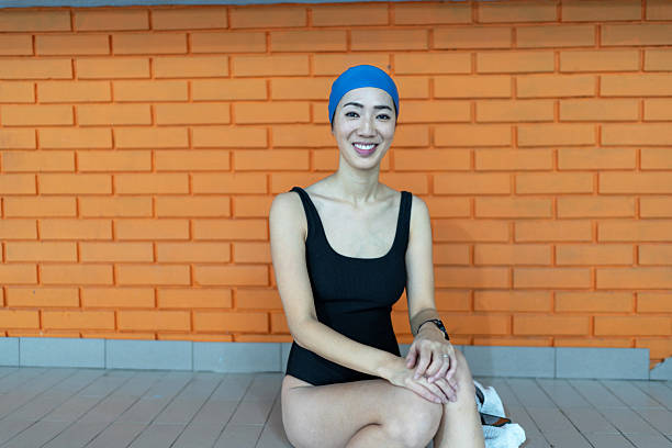 Smiling young adult asian woman sitting poolside, wearing a black swimsuit and a blue swimming cap, getting ready for a refreshing swim at an indoor pool