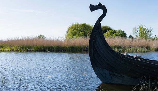 Close-up of the distinctive bow of a dark wooden Viking ship replica, featuring a carved dragon or serpent head prow. The ancient vessel floats peacefully on calm, rippling blue water, surrounded by a natural landscape of tall reeds and green trees under a bright, clear sky. The scene evokes history, exploration, and a serene, untouched natural environment.