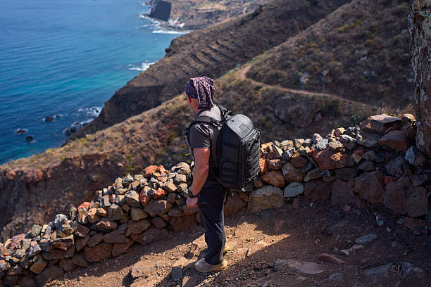 Male hiker with backpack and action camera stands by stone wall overlooking dramatic Atlantic coastal cliffs and terraced hillsides in Anaga Tenerife