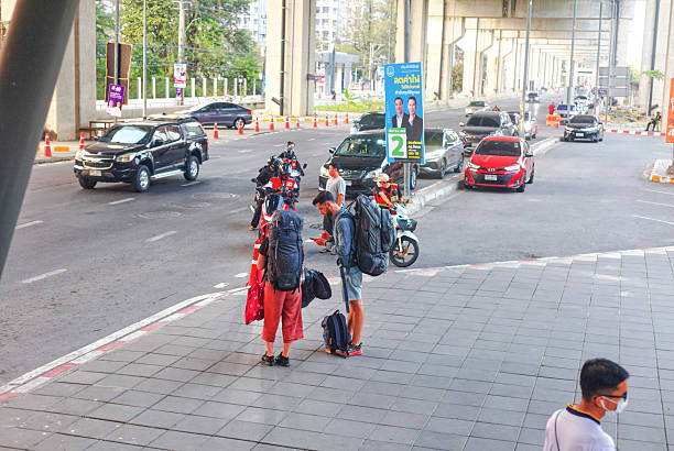 Young backpacker tourist couple below BTS Skytrain station  Don  Muang in Bangkok. People are having large backpacks. Man is using mobile phone and has a beard. They  are wearing  casual  clothes. People  are coming from Don Muang Airport via footbridge and down to street Vibhavadi Rangsit Road. In  background  are cars and some traffic. In right bottom area a person is walking with protectuve  face mask. Behind couple are some thai motorcycle taxi drivers