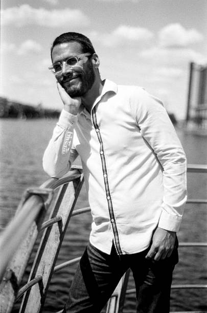 Black and white, grainy analog photograph of a bearded man in glasses, wearing a long-sleeved white shirt and dark pants, leaning on a metal railing by a river on a cloudy day