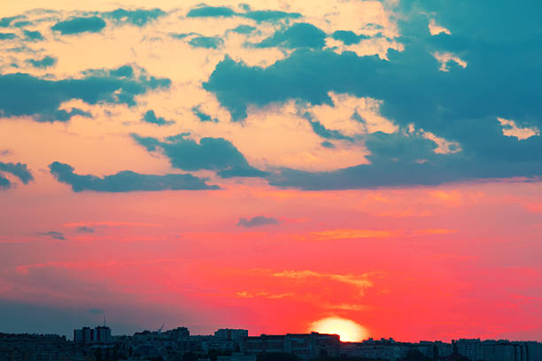 Sunset bathes city skyline in warm reds, oranges, and purples, silhouetting buildings against a dramatic cloud-filled sky. Vibrant gradient and moody atmosphere evoke awe and tranquility