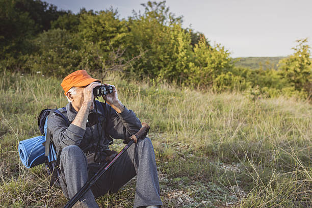 Senior birdwatcher wearing backpack is enjoying his retirement by hiking through nature, using trekking poles and binoculars looking for rare birds.