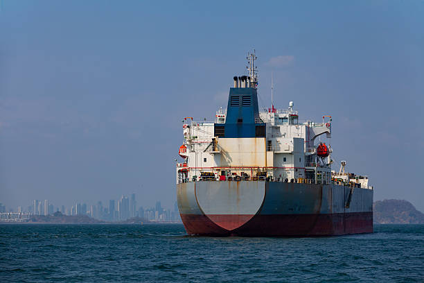 Large cargo ship viewed from the stern while anchored offshore and waiting to enter the Panama Canal. Calm sea conditions with a distant city skyline in the background illustrate global maritime transport, logistics, and international shipping routes in daylight.