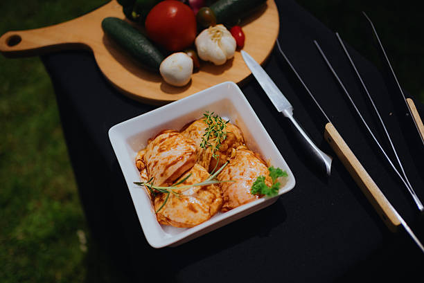 Bowl of raw, marinated chicken thighs with thyme and rosemary, beside fresh vegetables and grilling tools