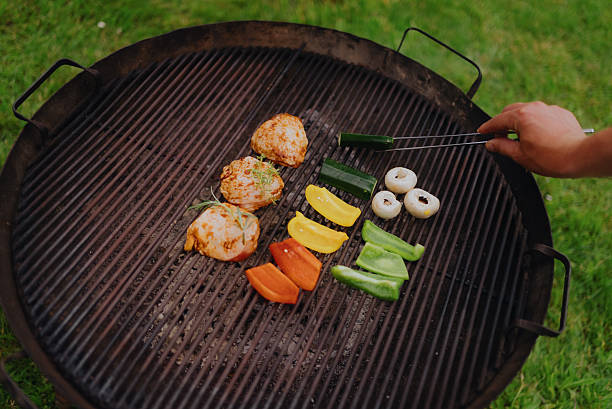 Hand using metal tongs for cooking healthy food, chicken thighs, zucchini, bell peppers, and mushrooms on a grill grate