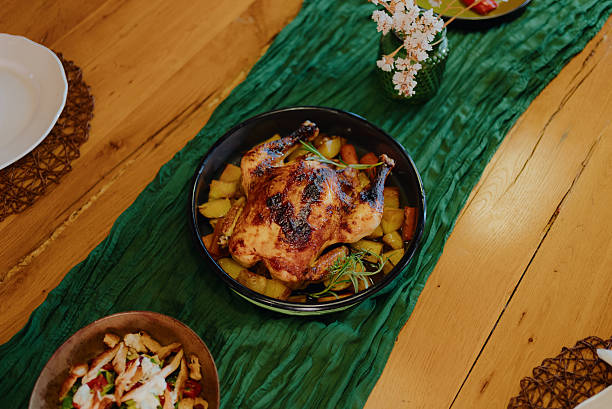 Whole roasted chicken and potatoes with vegetables, garnished with rosemary, sitting on a wooden dining table with a green runner