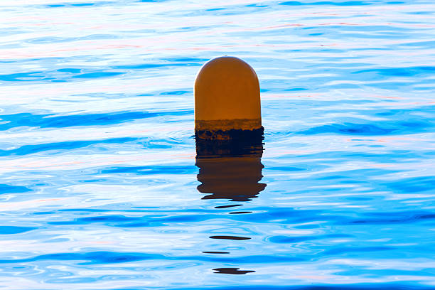 Single buoy floats in vibrant blue water. Sunlight highlights the gentle ripples. Water surface exhibiting pronounced, linear reflections and varying shades of azure surrounding floating safety marker