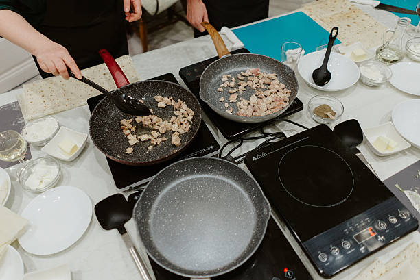 People learning to cook, frying chopped meat in pans on multiple induction cooktops during an organized culinary masterclass event