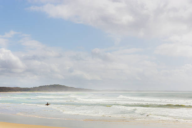 A lone surfer venturing with surfboard in hand wades out to the surf at Noosa Heads Beach, Sunshine Coast, Queensland, Australia.