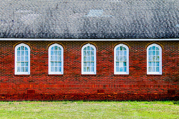 Church tall arched windows with white trim are set in a red brick wall below a weathered shingle roof and above a patch of green grass in a peaceful scene