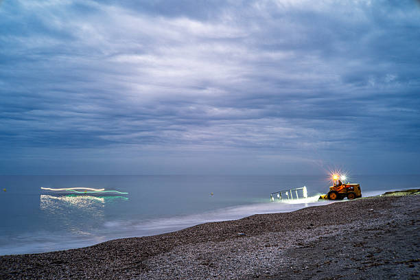 A tractor waits with trailer at dusk to remove a boat, indicated by light trails, from the water at Castell de Ferro, Granada, Spain. Long exposure.