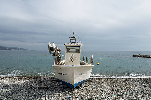 A white fishing boat, named Cala Chica, rests on a rocky beach near Castell de Ferro. The boat features a drawing of Virgin and Child on its front.