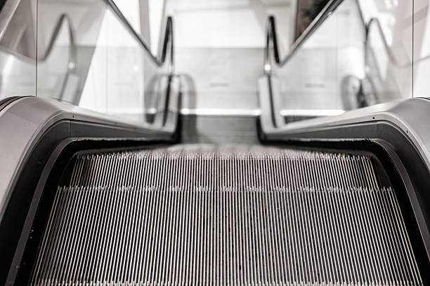Close-up view of an empty escalator going down, captured from the top step with a focus on the metallic grooves and glass side panels. Indoor, copy space