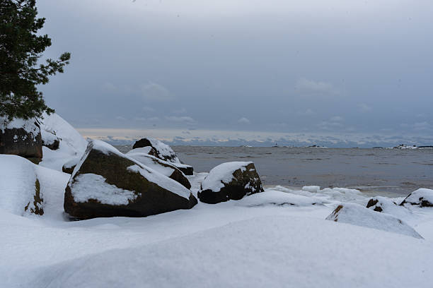 A serene winter landscape featuring snow-dusted boulders along a coastline, with choppy water and a hazy horizon dotted with islands