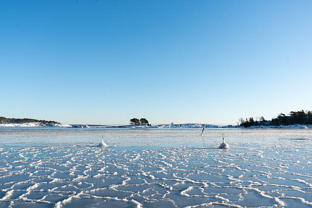 A vast expanse of frozen sea showcases delicate ice formations and navigational buoys, set against a backdrop of snow-dusted islands and a brilliant azure sky
