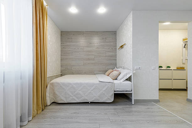 Horizontal view of a stylish studio apartment. Features a white metal bed with light quilted bedding, a grey wood-texture accent wall, and an entryway leading to a hallway with a white shoe cabinet.
