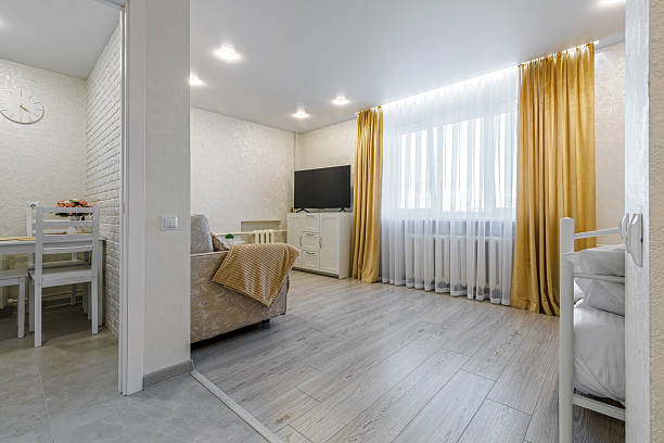 Wide horizontal view of a modern studio flat. Showcases a bright living room with a beige sofa, a TV area, and a glimpse into the dining nook. Large window with yellow curtains and light grey wood-style flooring.
