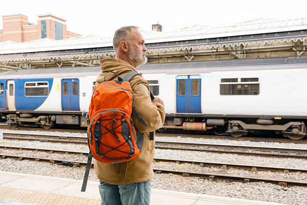 A man stands at a train station with an orange backpack. He is looking down the tracks as he waits for his train. The weather is cloudy and overcast.
