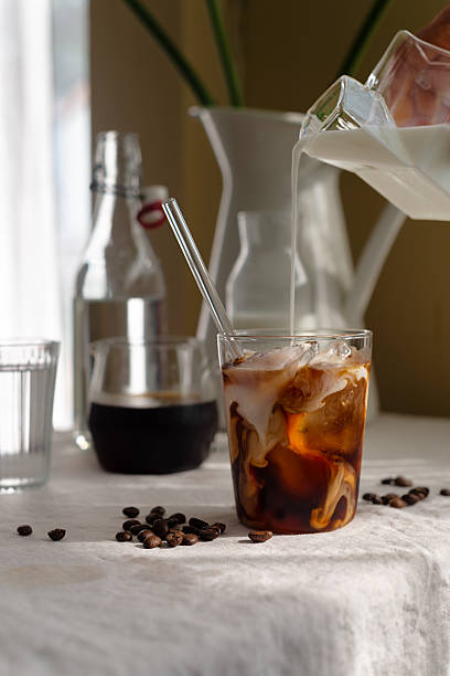 Pouring milk into a glass of homemade cold brew coffee on white table background.