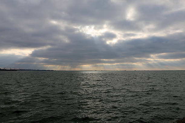 The sea with dark cloudy skies and sun rays shining through them. The coast and harbor are visible in the distance.