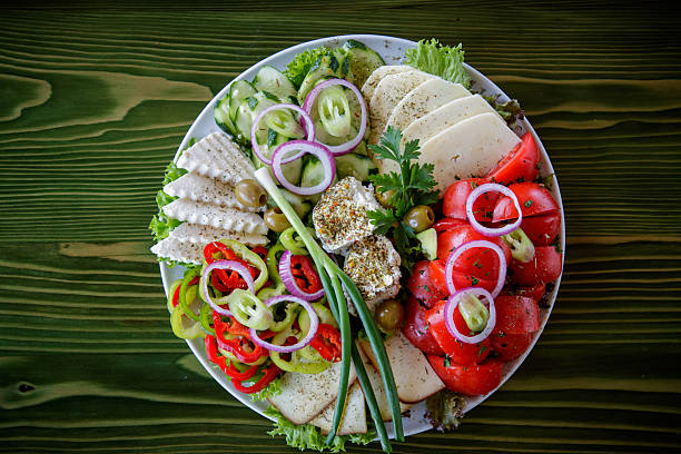 Sliced pepper, onion, tomato, cucumber, lettuce leaves, scallion, green olives and three types of cheese on appetizer plate on restaurant table, garnished with parsley and basil, directly above