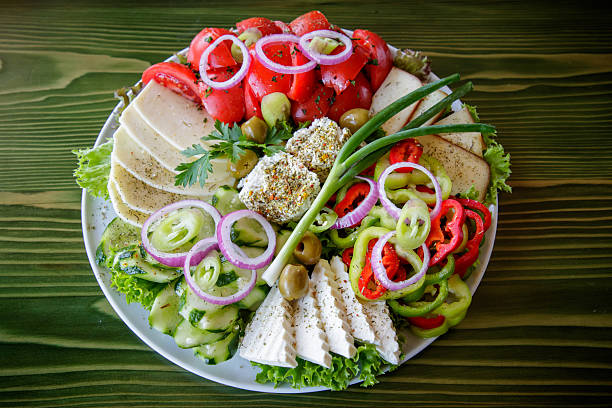 Sliced pepper, onion, tomato, cucumber, lettuce leaves, scallion, green olives and three types of cheese on appetizer plate on restaurant table, garnished with parsley and basil
