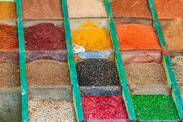 Close-up of a market stall showing a grid of compartments filled with mounded spices and dried seasonings. Green dividers separate piles of powders, seeds and flakes in vivid yellows, oranges, reds, blacks, pinks, greens and neutrals, highlighting varied textures, colors and abundance typical of a spice vendor.