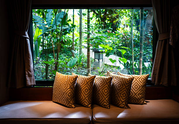 Window corner decorated with square pillows on sofa with blurry background of tropical garden.