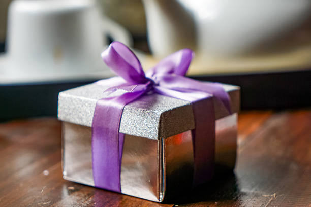 Shiny silver gift box with purple ribbon on wood table, teapot and flowers in soft background