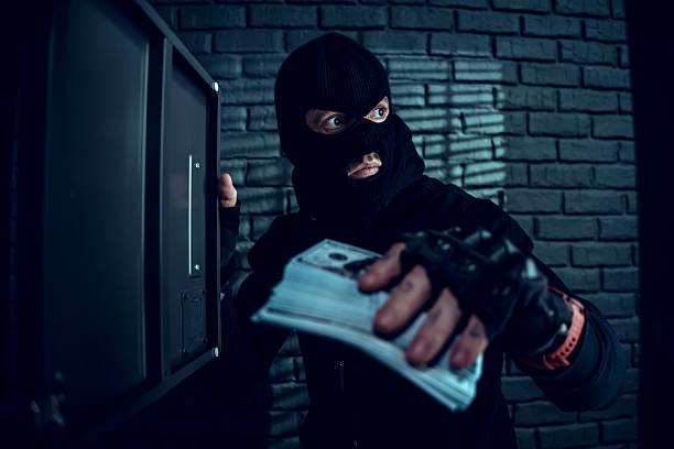 Criminal in black balaclava holding money bags while robbing a safe in a dark room with brick walls.