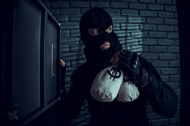Criminal in black balaclava holding money bags while robbing a safe in a dark room with brick walls.