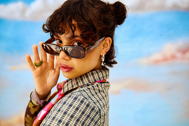 Young woman raising sunglasses against a blue sky backdrop, looking at camera