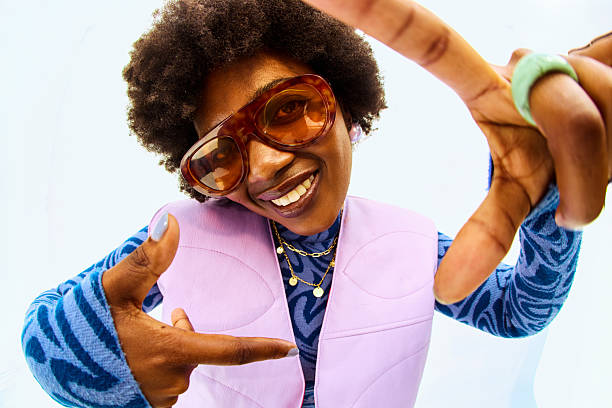 Smiling young adult woman framing face with hands, wearing retro fashion and sunglasses in a studio
