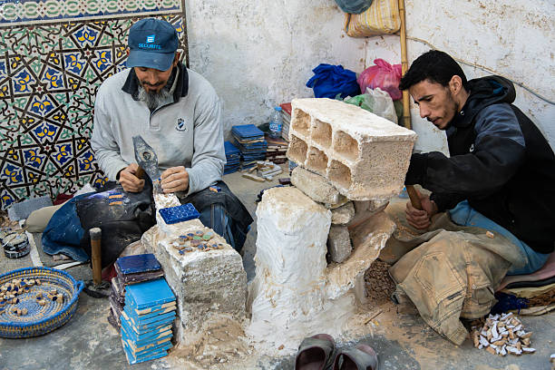 Two craftsmen shape clay and stack bricks in a Fez pottery workshop, surrounded by tiles, tools, and raw materials. A scene of traditional craft and focused labor in Morocco.