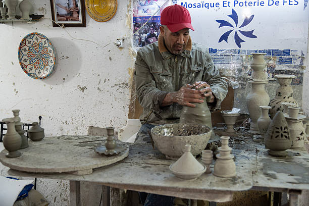 A Moroccan potter shapes clay on a dusty table in a Fes workshop, surrounded by finished and in-progress pieces. The scene radiates craftsman focus and traditional pottery artistry in Morocco.