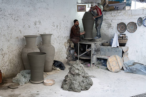 A Moroccan pottery studio in Fez featuring two potters shaping a tall vase, surrounded by large kilned vessels, plates on walls, and a rugged worktable. Earthy tones convey craft and tradition.