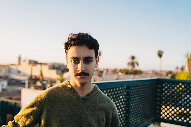 Young adult man with mustache looking at camera, standing on a rooftop at sunset with a cityscape in Marrakech
