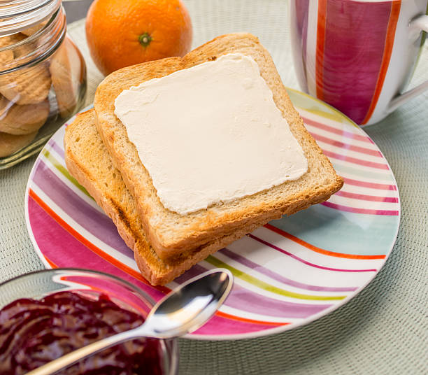 A woman is seen spreading butter on a slice of bread while preparing breakfast in the kitchen. She is using a spoon to apply butter from a small bowl, with various fruits like oranges and a pear visible on the table along with a knife.