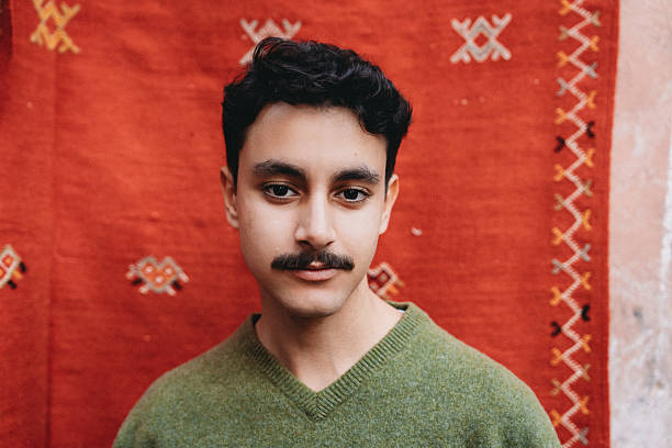 Young adult man with mustache looking at camera, standing in front of a red traditional Moroccan Berber rug