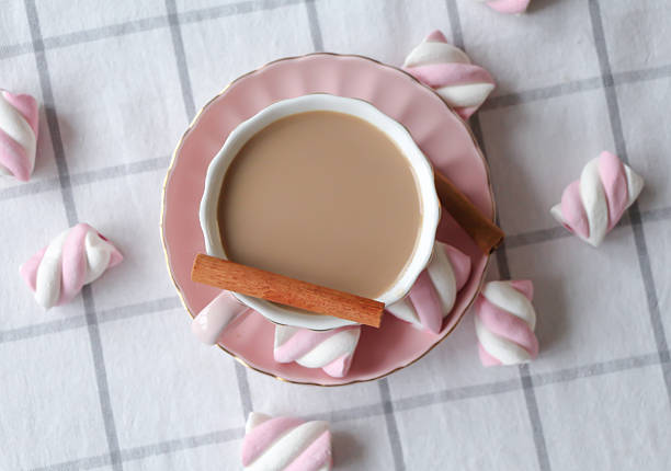 Top view flat lay of a pink cup with milky tea and cinnamon on a matching saucer surrounded by pink-white marshmallows on a light checkered textile background. Minimal cozy concept with copy space.
