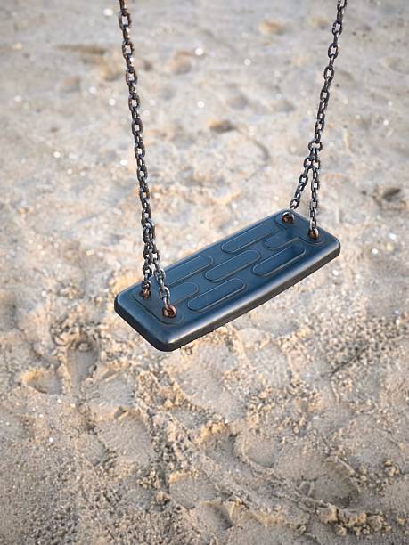 A close-up of an empty swing seat on metal chains hanging over sand at a children's playground. Conceptually suggesting loneliness or waiting.