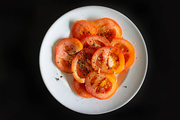 Top view of fresh sliced tomatoes drizzled with olive oil and seasoned with black pepper, served on a white plate against a black background.