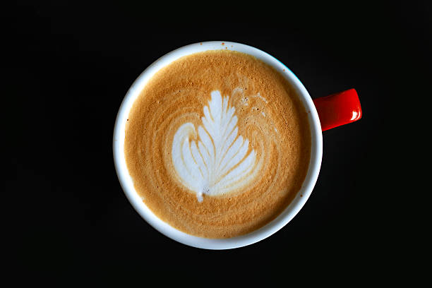 Close-up, top view of a latte coffee featuring delicate milk foam art served in a ceramic cup against a black background.