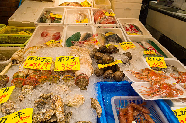 Osaka, Japan - November 7, 2023: A seafood stall at Kuromon Ichiba Market displays scallops, clams, oysters, shrimp, and various fish on ice in trays and styrofoam boxes, with handwritten price signs in Japanese visible.