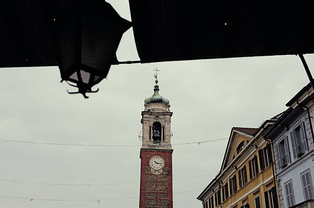 Historic bell tower with a clock rising above traditional urban buildings on an overcast day. The tower is framed by surrounding architecture and foreground elements, creating a balanced cityscape composition. Concept of time, history, European city life, and architectural heritage. No people visible.