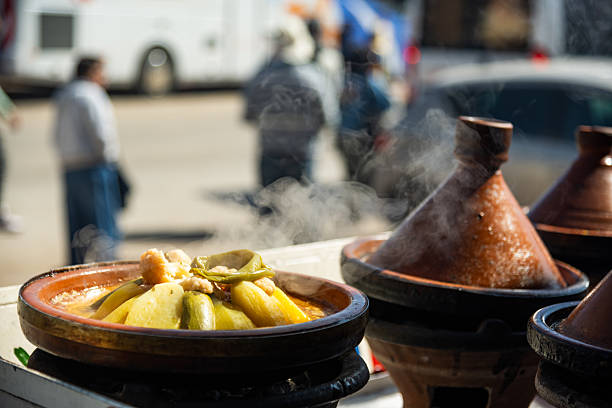 A steaming Moroccan tagine filled with colorful vegetables and meat sits on a traditional clay pot, while blurred pedestrians pass in the background. Perfect for food, street cuisine, and cultural dining themes.