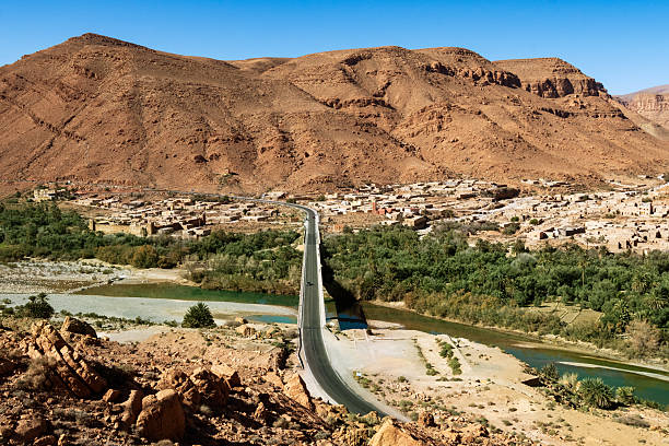 A arid Moroccan desert town sits beneath rugged hills, crossed by a long bridge over a winding river. Lush greenery and palm trees contrast with the red mountain backdrop.